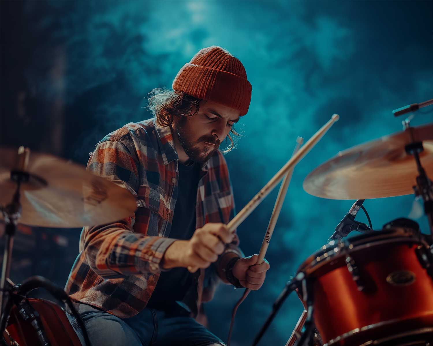 A man wearing a red beanie and plaid shirt plays a drum set on stage, with blue smoke in the background.